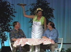 A woman, holding a waitress tray, talks with two men sitting at a table with a checkered table cloth.