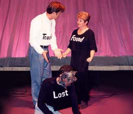 An image from the play, "God, Look!" in which the "Teacher" motions to "Lost," while "Found" looks on. 