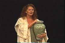 A woman, dressed in Bible costume, holds a large green water vase.