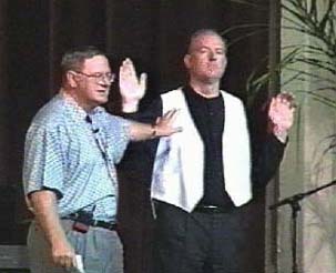 A man in a blue shirt points to a man with a vest on.