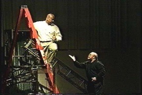 A man on a ladder listens to a man gesturing on the floor below him.