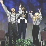 A woman holds a book and two mimes in striped shirts leap up with their hands raised.