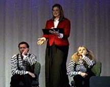 A woman holds a book, and gestures to two mimes in striped shirts.